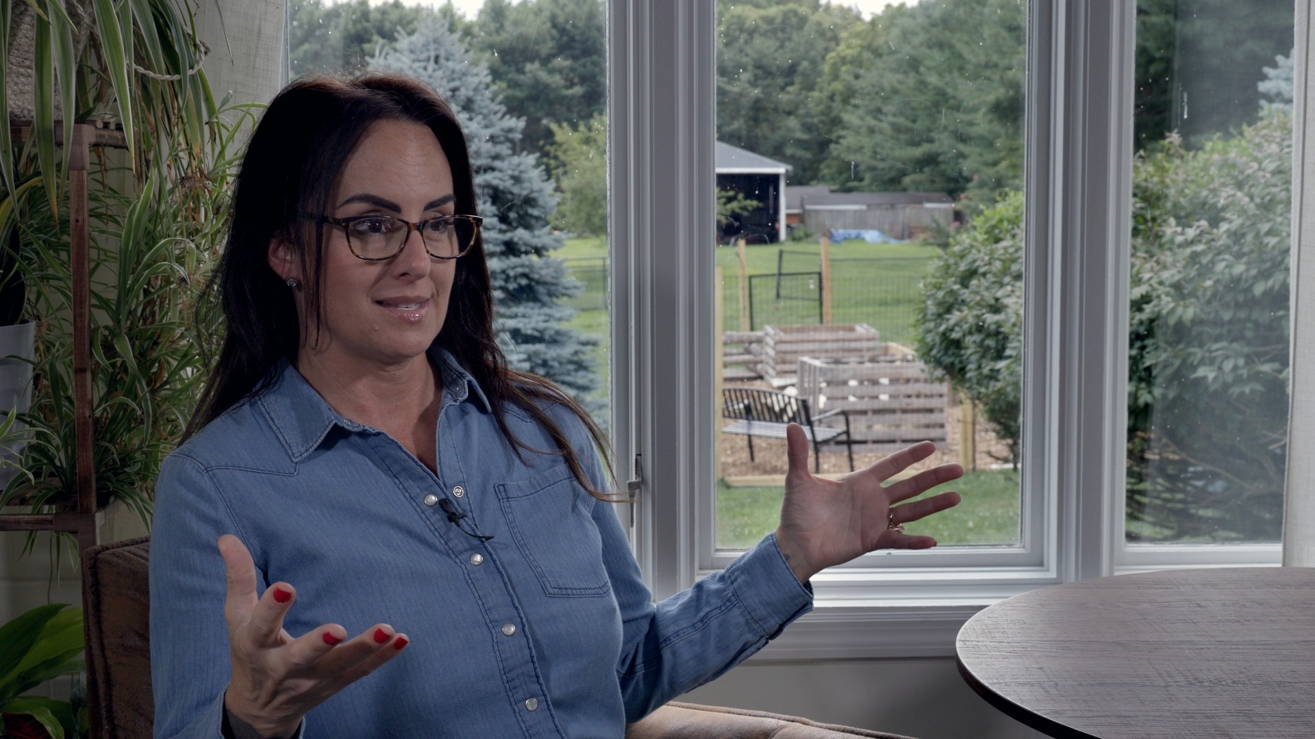 A woman in a denim shirt speaking during an interview beside a bright garden window